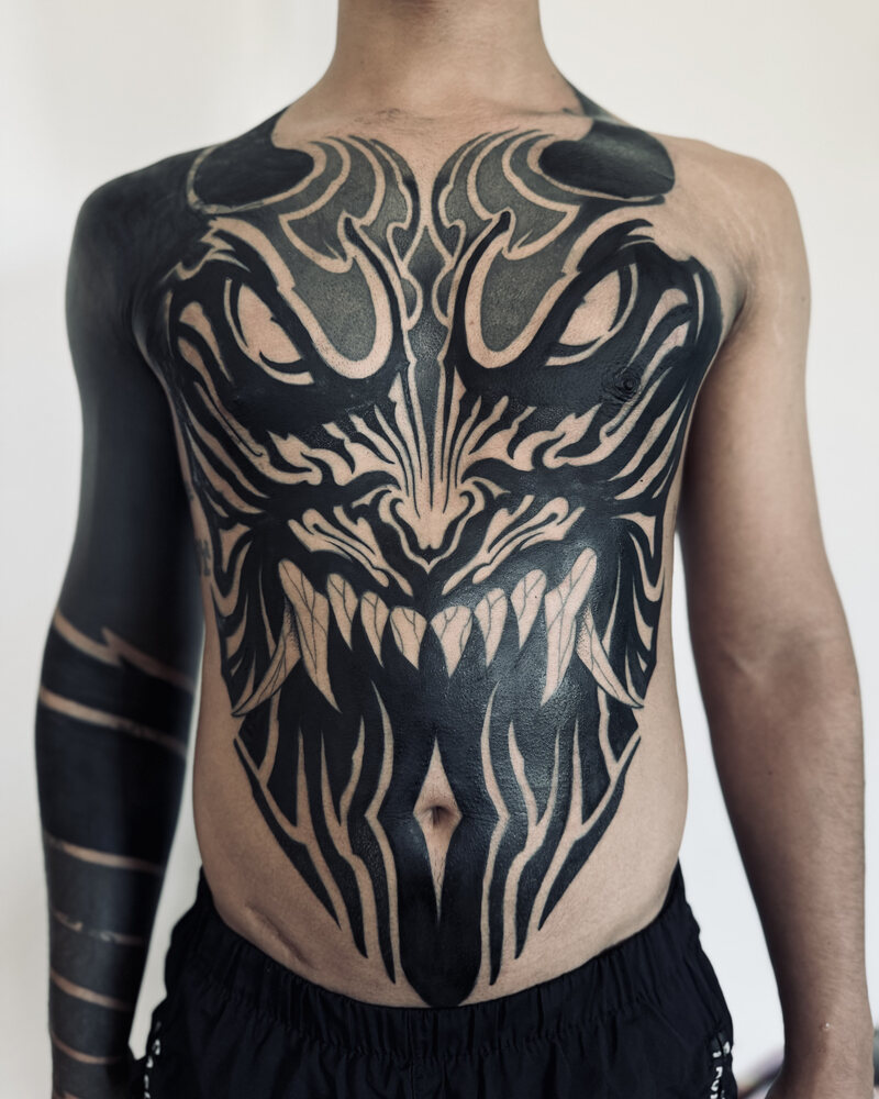 Tatuaje blackout de una criatura con colmillos en pecho y abdomen, con el brazo en negro sólido