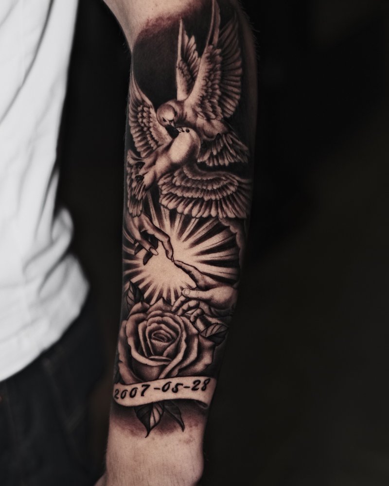 Tatuaje en negro y gris: ángel con rosas y una banda con la fecha 1987-06-21, en el antebrazo