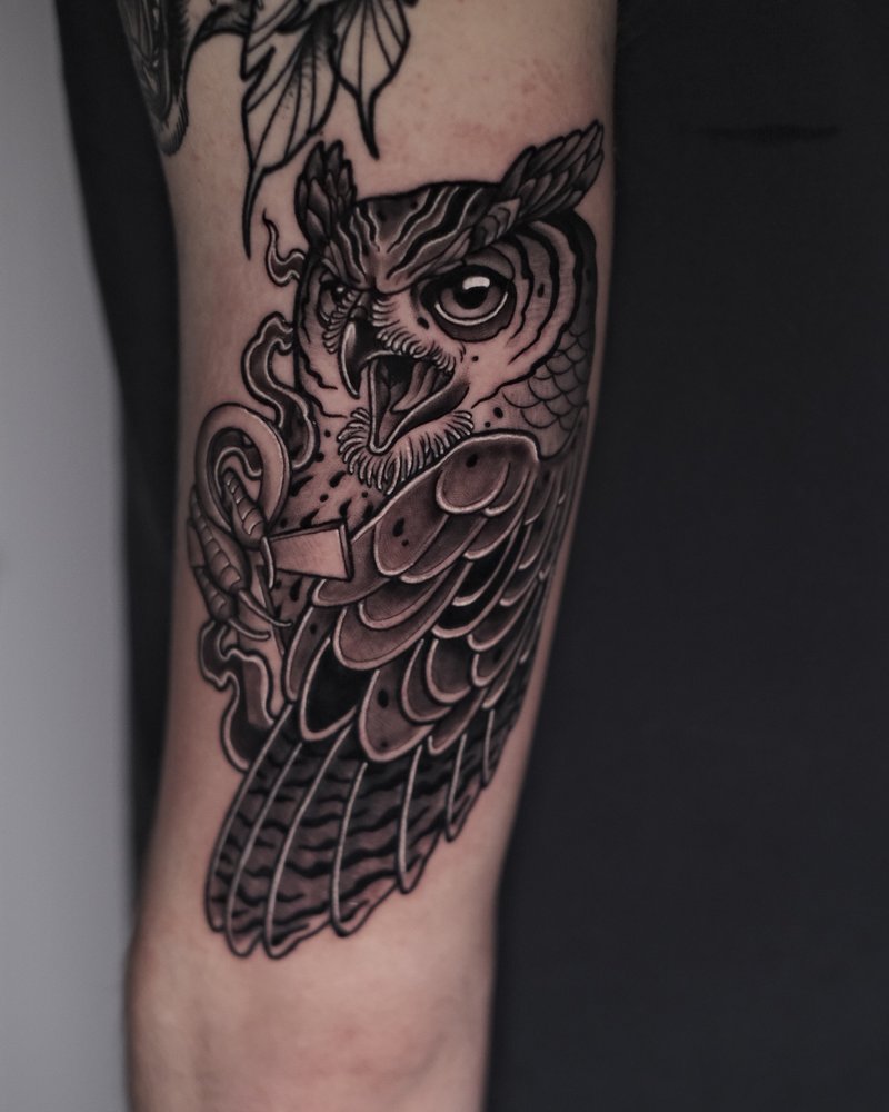 Tatuaje en negro y gris de un búho con ornamentos, en el antebrazo