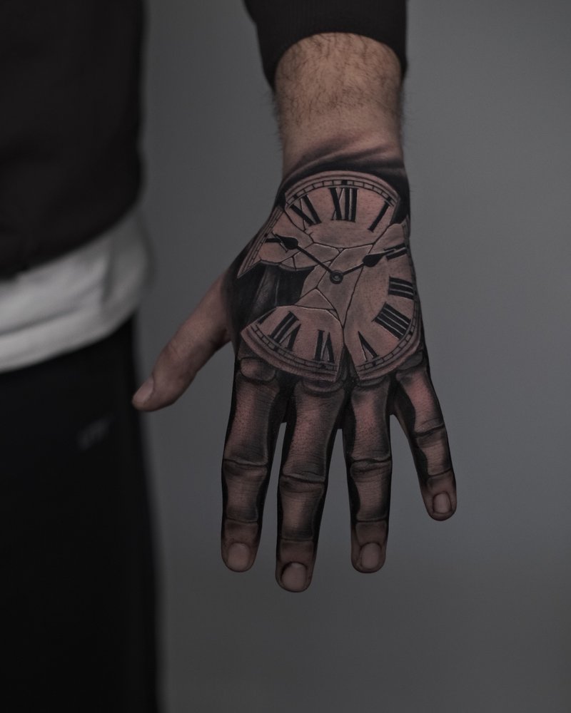 Tatuaje de un reloj en negro y gris en el dorso de la mano