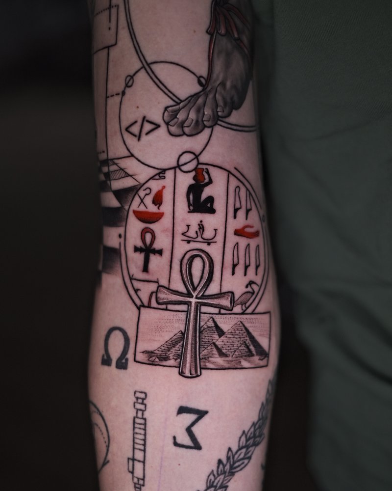 Tatuaje de línea fina con un ankh, un reloj de arena y un ojo, en el antebrazo