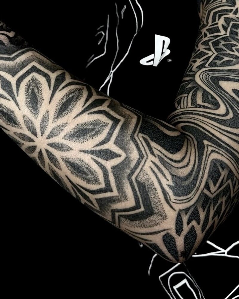Tatuaje de manga con mandala floral geométrico y líneas onduladas en el brazo, en negro