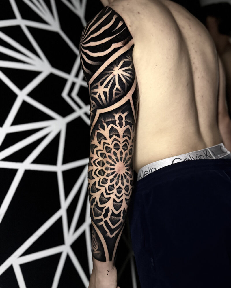Tatuaje de manga geométrica con mandala en el antebrazo y patrones de estrellas en el codo, en negro
