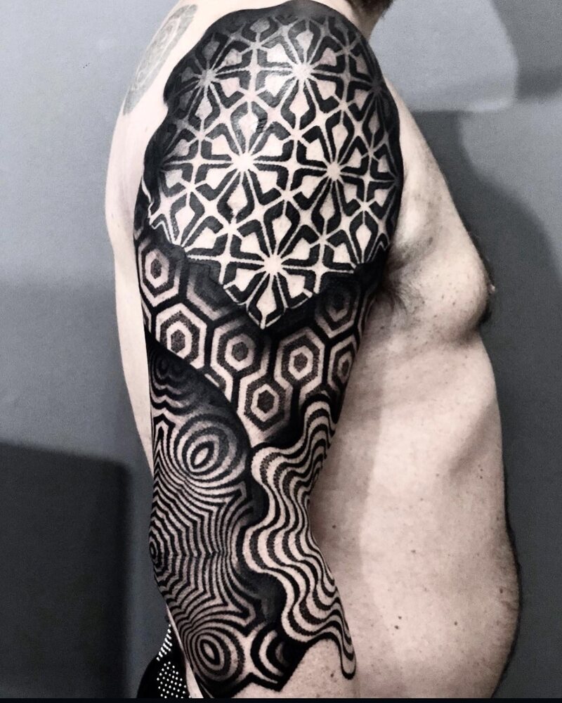Tatuaje de geometría en hombro y brazo con patrón de estrellas, hexágonos y líneas concéntricas, en negro