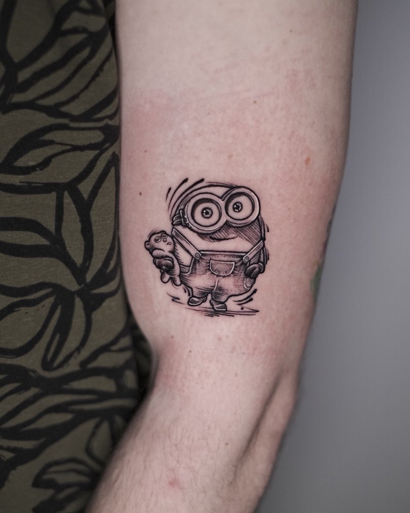 Tatuaje en negro y gris de un minion con un gato, en el brazo