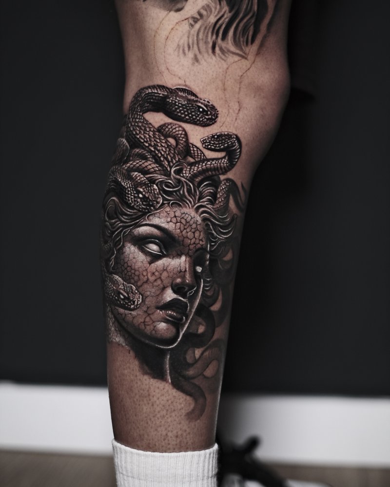 Tatuaje realista en negro y gris de un rostro de medusa con serpientes, en la pierna
