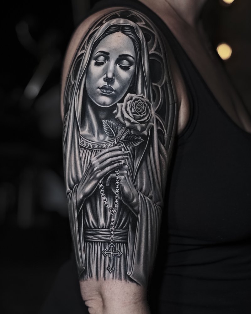 Tatuaje en negro y gris de una Virgen María con un rosario, en el brazo
